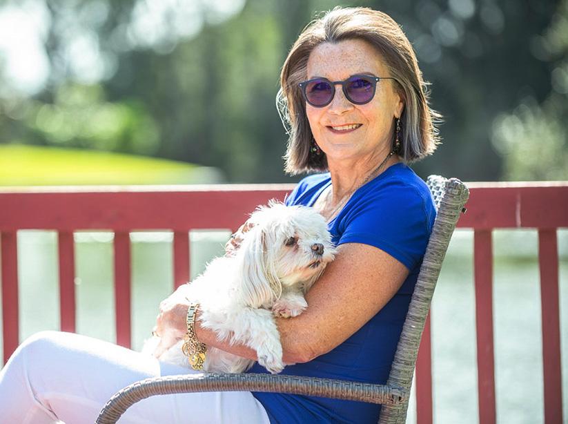 A woman wearing purple-tinted sunglasses, a blue shirt, and white pants sits outside, holding a small white dog, and smiles at the camera. 