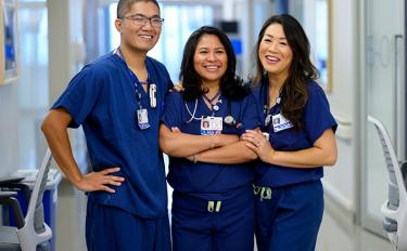 A man with close-cropped hair stands in a hospital hallway with two women with long dark hair, all wearing blue hospital scrubs and smiling.