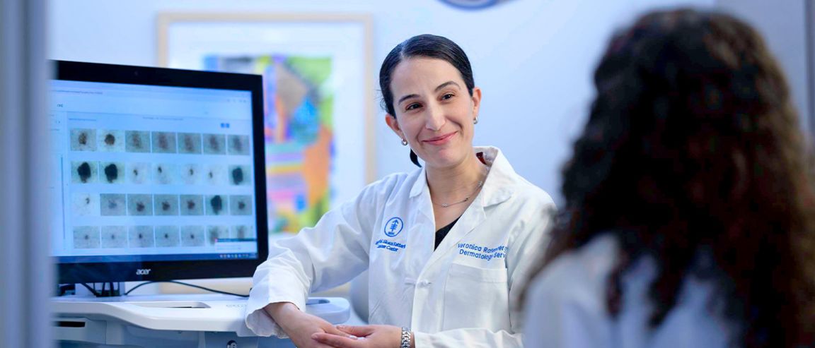 MSK’s Veronica Rotemberg MD, PhD, smiles while in conversation with another person. A monitor showing images of skin cancer is behind her.
